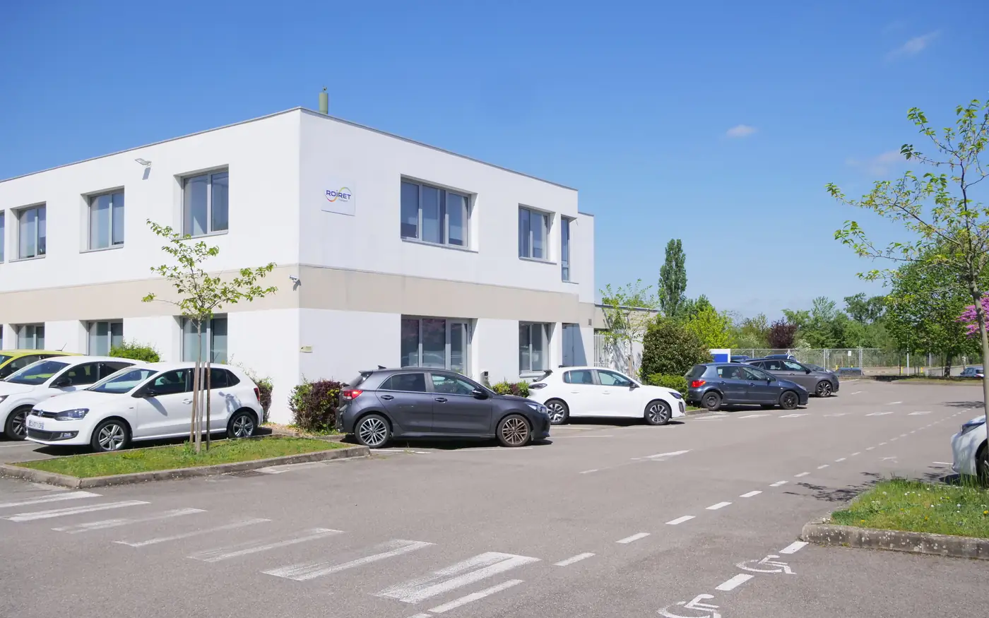 Vue au niveau du sol du parking d'ENFRASYS avant travaux : voitures alignées devant un bâtiment blanc, marquage au sol sur enrobé.