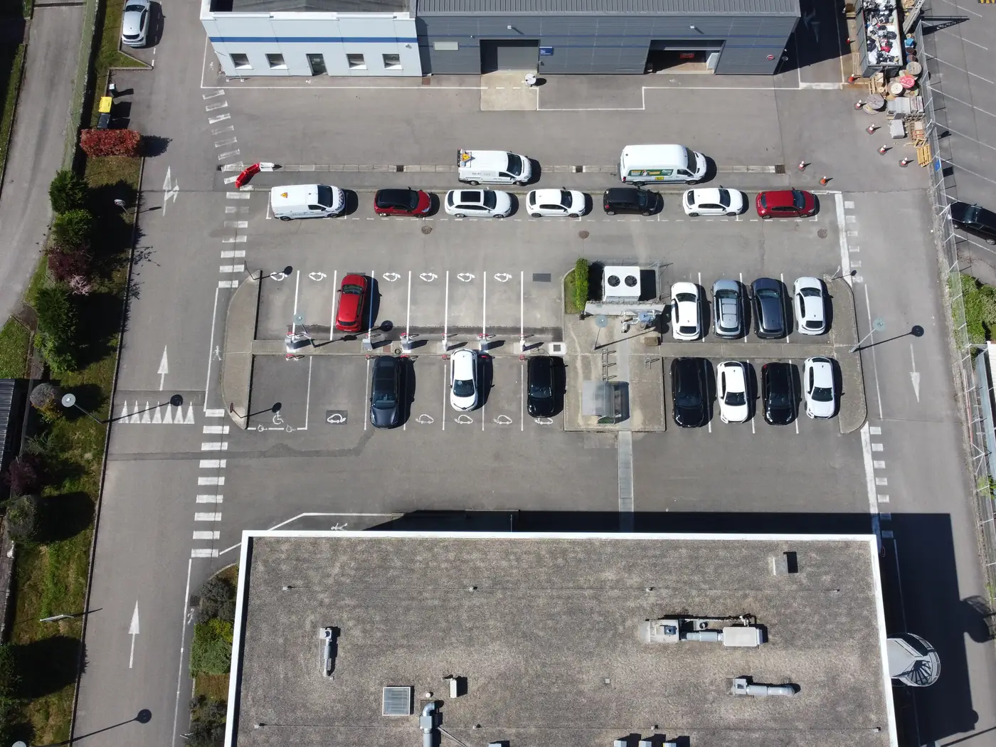 Vue verticale au drone du parking d'ENFRASYS : rangées de voitures sur un enrobé continu, sans aucun arbre.